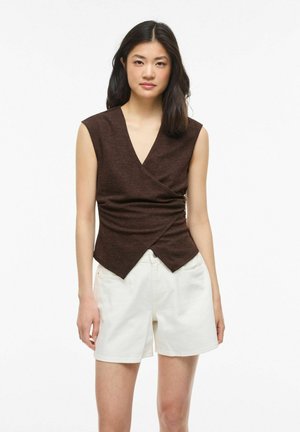 Młoda kobieta z ciemnymi włosami do ramion nosi ciemnobrązową, bezrękawną bluzkę kopertową i białe szorty do kolan, stoi na białym tle.