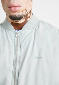 Gaiši zila bomber jaka, kas izgatavota no gludas auduma, ar priekšējo rāvējslēdzēju un izšūtu "OBEY" logo uz krūškurvja. Redzama silikona tekstūra.