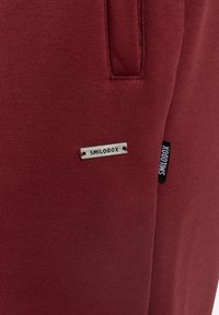 Detailní záběr na vínový materiál s přední kapsou a dvěma štítky značky "SMILODOX", jedním kovovým a jedním černým textilním.