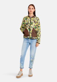 Blumige gesteppte Jacke in Grün, Gelb und Braun, mit Taschen und Reißverschluss. Kombiniert mit hellblauen Jeans mit floralen Stickereien.