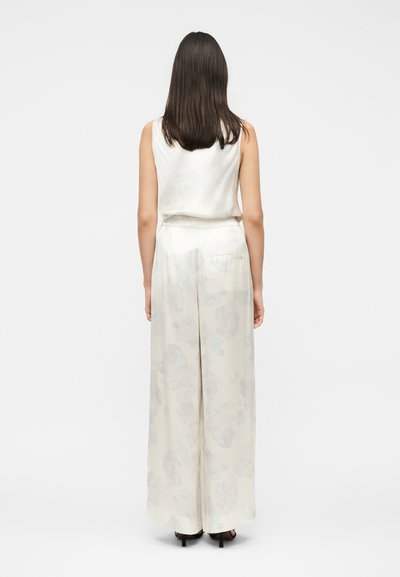 Vrouw met donker schouderlang haar die een mouwloos wit topje en een witte, hoog getailleerde broek met wijde pijpen draagt, staat met haar rug naar de kijker.