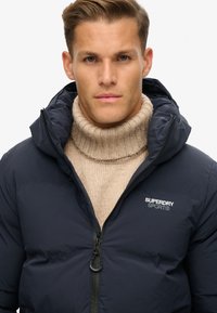 Marineblå puffjakke med lynlås fortil, justerbar hætte og et sort logo på brystet. Båret over en beige strikket højhalset sweater.