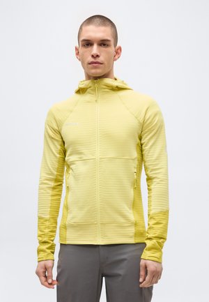 Młody mężczyzna z krótkimi włosami, ubrany w jasnżółtą kurtkę z kapturem i szare spodnie, stoi na tle jednolitego jasnego tła.