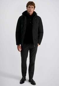 Zwarte pufferjack met capuchon en voorzakken, gedragen over een zwarte trui. Grijze gemêleerde broek en zwarte enkellaarsjes maken de look compleet.