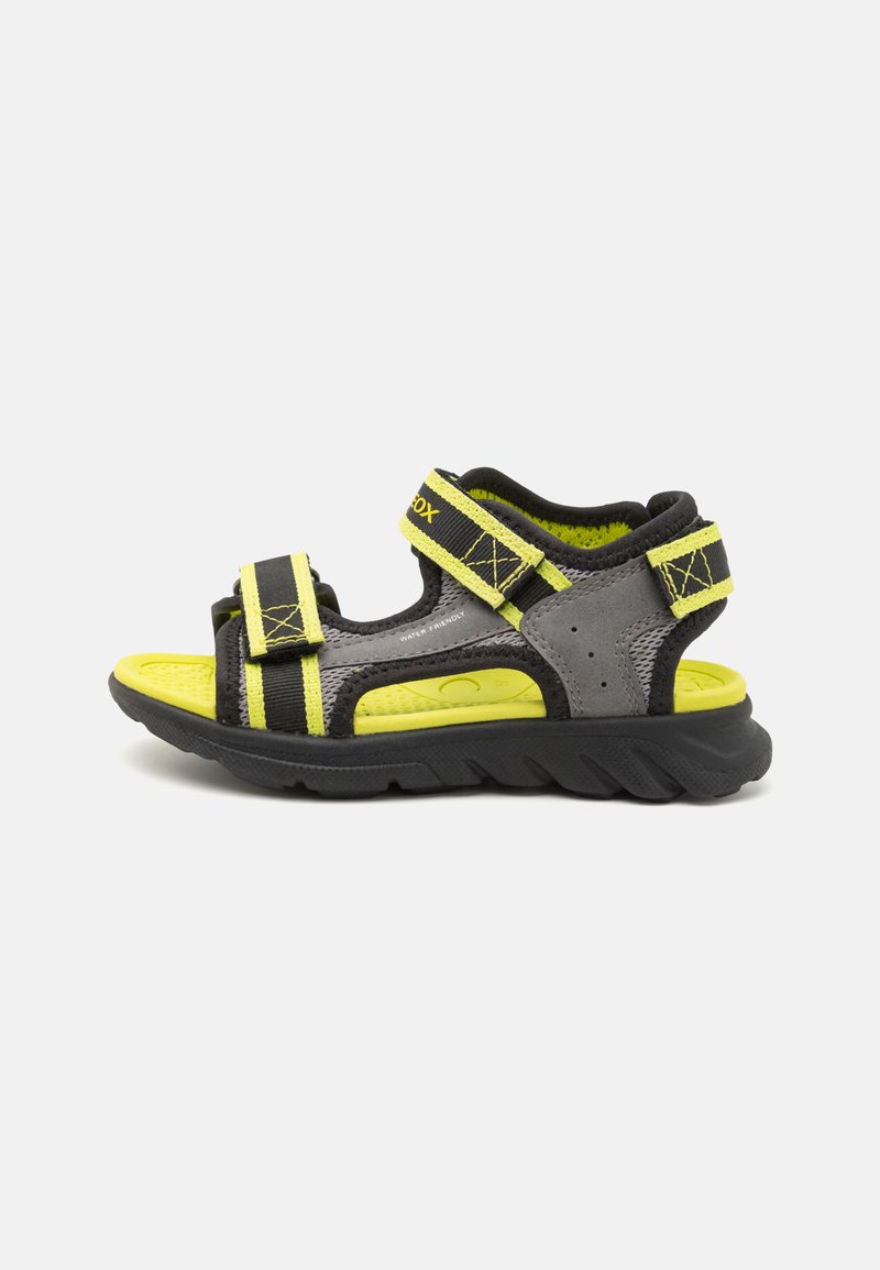 Svart och grå sandal med gula detaljer, utrustad med justerbara remmar, en strukturerad sula och en flexibel, robust sula. Vattenvänlig design.