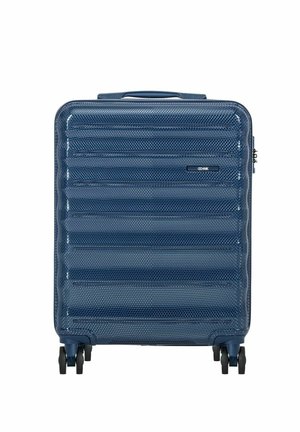 Niebieska teksturowana walizka z twardą skorupą, wyposażona w cztery czarne obrotowe kółka i górny uchwyt do przenoszenia, stojąca pionowo na białym tle.