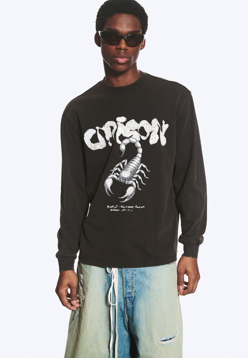 Jeune homme portant une chemise noire à manches longues avec un graphisme de scorpion blanc et un jean bleu clair déchiré, associé à des lunettes de soleil noires.