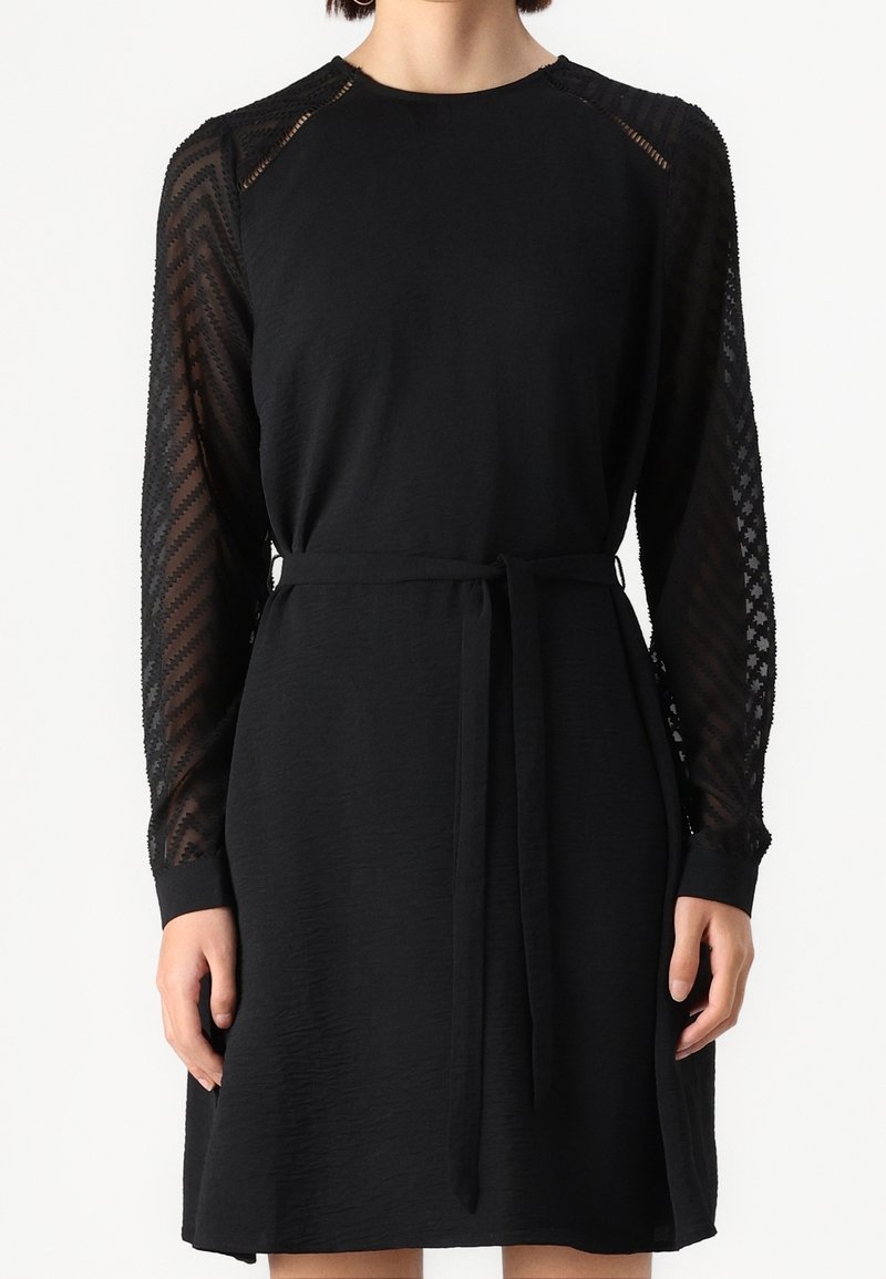 Femme portant une robe noire longueur genou avec des manches longues transparentes à motifs et une ceinture en tissu nouée à la taille.