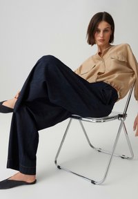 Femme aux cheveux courts et bruns, assise de côté sur une chaise pliante en métal, portant un chemisier beige, un pantalon large foncé et des chaussures noires sans lacets.