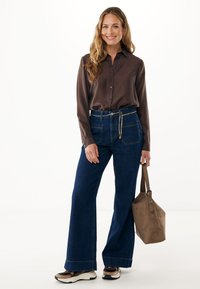 Camicia button-up marrone scuro, pantaloni a gamba larga in denim blu a vita alta e borsa in suede beige con accento a catena. Sneakers con design nero e beige.