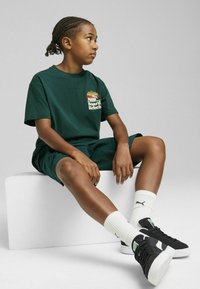 Conjunto de camiseta de algodón verde y pantalones cortos que presenta un logo en amarillo y rojo, complementado por calcetines acanalados blancos y zapatillas negras.