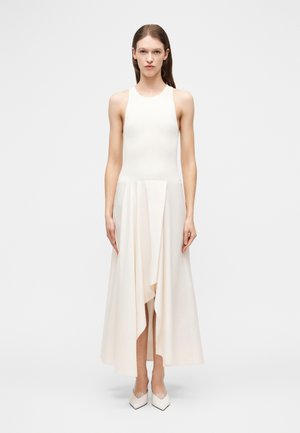 Femme portant une robe midi asymétrique sans manches de couleur crème et des escarpins blancs à bouts pointus, debout devant un fond blanc uni.