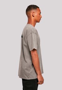 Young man with short curly hair stands in profile wearing a loose, faded gray T-shirt and black pants against a plain light background.