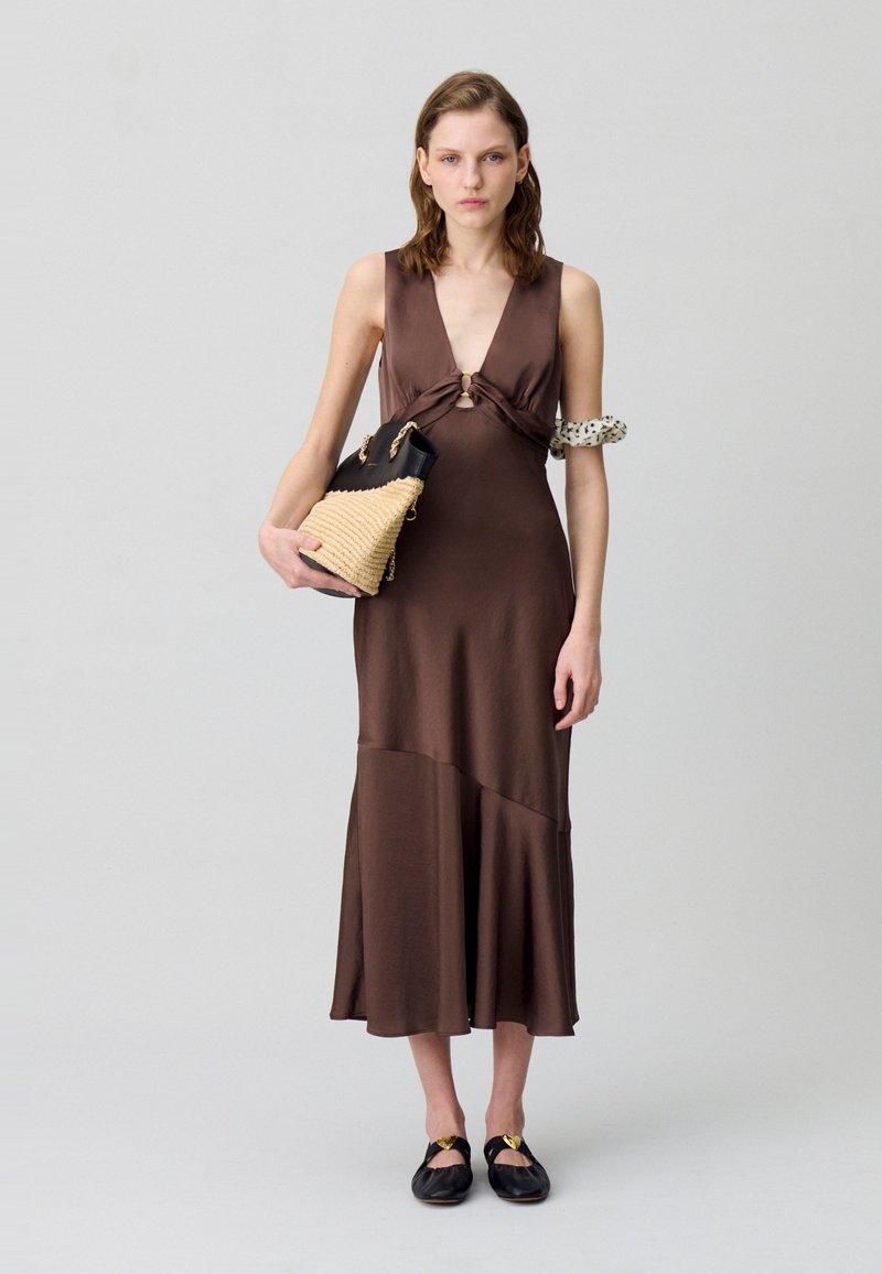 Robe sans manches marron avec un détail de nœud torsadé et un décolleté en V. Un sac à main noir tressé présente des accents dorés. Le mannequin porte des chaussures noires.