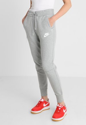 Osoba ubrana w szare spodnie dresowe Nike i jaskrawoczerwone buty Nike stoi z rękami w kieszeniach.