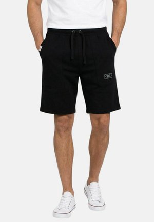 Homme portant un short noir mi-genou à taille ajustable, des baskets blanches et un t-shirt blanc, les mains dans les poches, debout sur un fond uni.