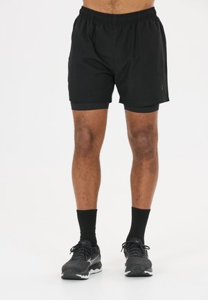 Homme portant un short de course noir avec un short de compression intégré, des chaussettes noires et des chaussures de course noires, debout sur un fond blanc.