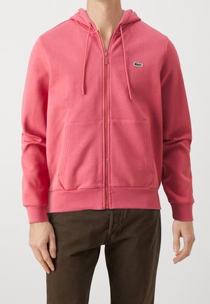Homme portant un sweat à capuche zippé rose avec un petit logo crocodile vert sur la poitrine et un pantalon marron sur un fond uni.