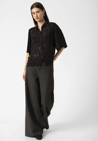 Vrouw in een zwarte doorzichtige bloemenblouse met halve mouwen en een wijde donkergrijze broek, staande tegen een effen lichte achtergrond.