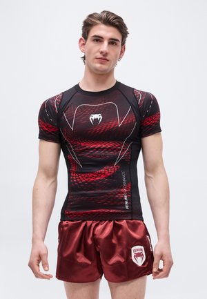 Noor mees, kes kannab tihedat musta ja punast maodermooni mustriga spordisärki ning läikivaid punaseid Venum spordipükse, seisab ühevärvilise tausta ees.