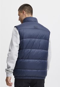 Tummansininen puhvivestsari, jossa on korkea kaulus, vaakasuora tikkauskuvio ja logotagsi takana. Päällä harmaa pitkähihaiseksi paita.