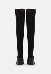 Botas de ante negras hasta la rodilla con tacones de bloque, vistas desde atrás, con cordones atados en la parte superior de cada caña.