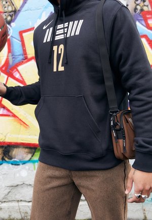 Sudadera negra de Nike con rayas verticales blancas y el número "12" en dorado. Combinada con pantalones marrones texturizados y un bolso cruzado marrón.