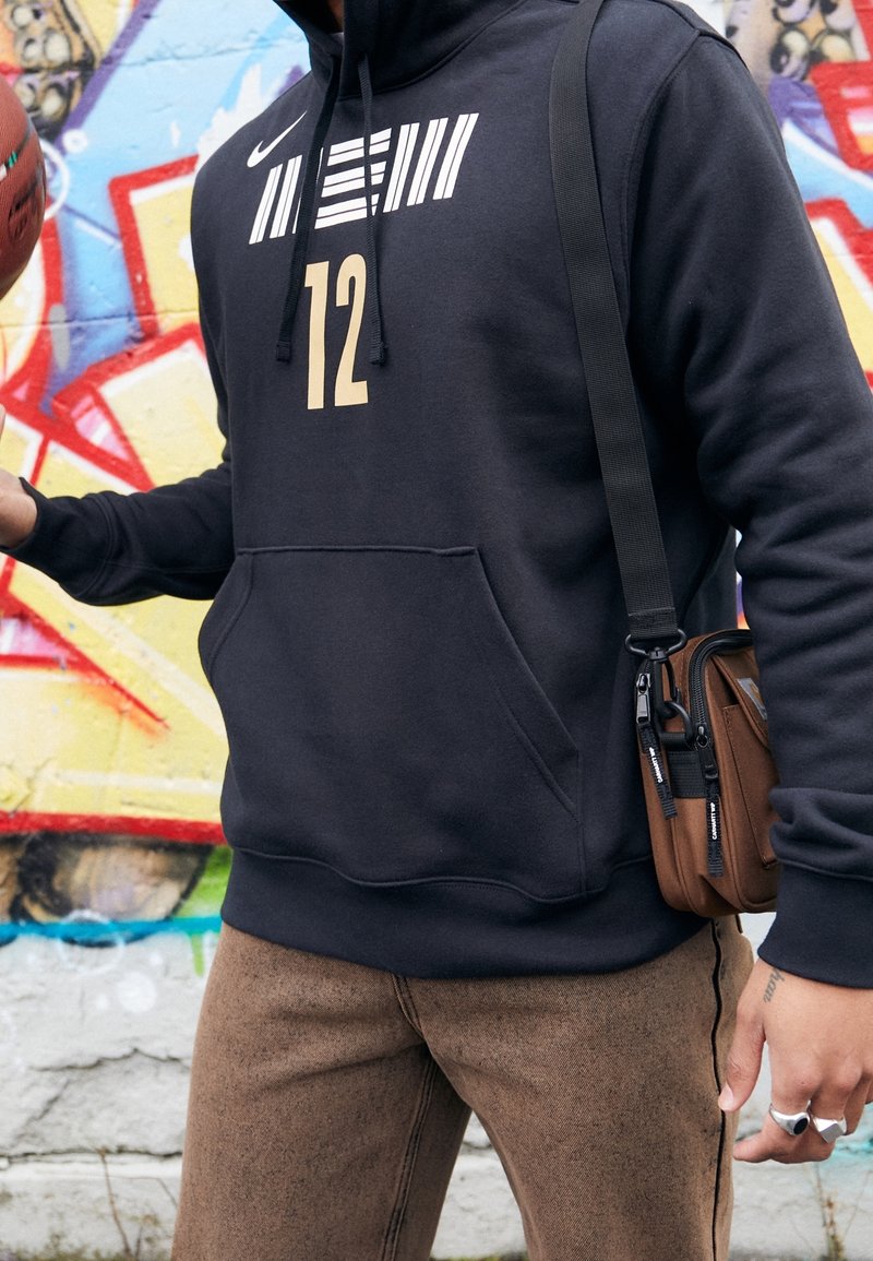 Felpa nera Nike con strisce verticali bianche e il numero "12" in oro. Abbinata a pantaloni brown con texture e una borsa a tracolla marrone.