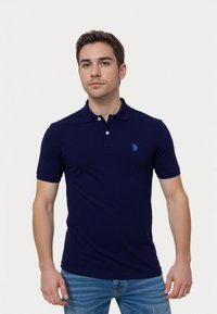 Young man in a navy polo shirt and blue jeans, standing with a neutral expression against a white background.