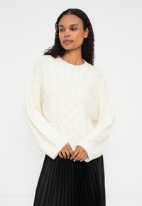 Suéter blanco de punto trenzado con cuello redondo, mangas oversized y puños acanalados, combinado con una falda negra de tablas. Tejido suave y texturizado.