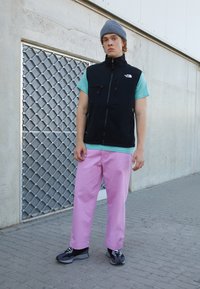 Juodas užtrauktuku užsegamas liemeniukas, turquoise spalvos marškiniai, rožinės kelnės ir juodi/pilki sportbačiai. Fone matoma tekstūruota pilka siena ir tinklinė tekstūra.