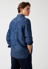 Denimskjorte i dybblå med indviklet mønsterstruktur, lange ærmer oprullet, og en buet søm bagpå. Bagfra.