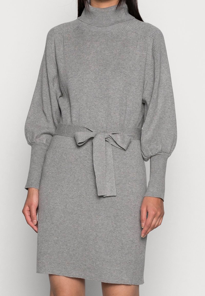 Femme portant une robe en maille côtelée grise avec col roulé, manches longues bouffantes et ceinture nouée à la taille.