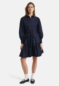 Rochie chemiză bleu marin cu front cu nasturi, mâneci lungi tip balon, curea la talie, și fustă evazată în straturi. Țesătură ușoară.