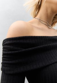 Czarny prążkowany top z odkrytymi ramionami i fakturą, ozdobiony warstwowym srebrnym naszyjnikiem spoczywającym na obojczyku.