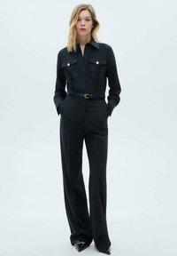 Svart jumpsuit med krage, två bröstfickor med guldknappar, midja med bälte och vida byxor, tillverkad av slät tyg.