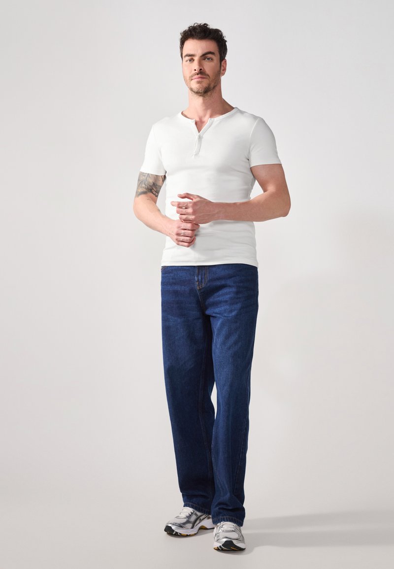 Witte korte mouw henley shirt met een knoopsluiting, gecombineerd met donkerblauwe recht gesneden denim jeans en grijze sportschoenen.