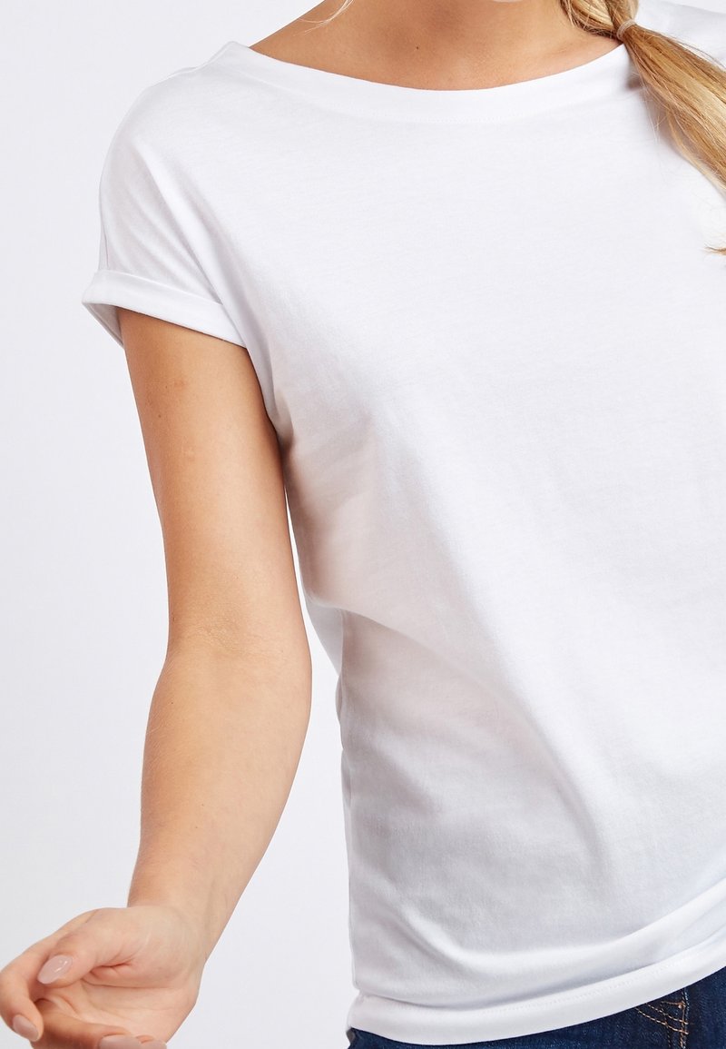 Femme portant un t-shirt blanc uni à manches courtes retroussées et un jean bleu, visible des épaules à la taille.