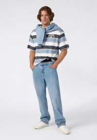 Jonge man die met handen in zijn zakken staat, gekleed in een gestreepte Champion T-shirt, lichtblauwe jeans, witte sneakers en een blauwe sweatshirt over zijn schouders.