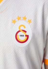 Jersey blanc texturé avec un motif en tourbillon subtil. Présente un logo circulaire avec des accents rouges et jaunes, quatre étoiles jaunes au-dessus.