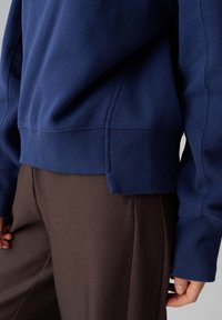 Marineblauer Pullover mit gerippten Bündchen und Saum, mit lockerem Schnitt und weicher Textur. Kombiniert mit brauner Hose, die minimale Nähte aufweist.