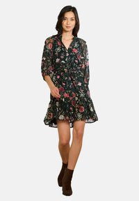 Kobieta w czarnej sukience w kwiaty z falbaniastym dołem i rękawami 3/4, w brązowych botkach za kostkę, idąca do przodu na białym tle.