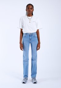 Camiseta blanca de manga corta con texto verde, combinada con jeans rectos de color azul claro y complementada con zapatillas metálicas.