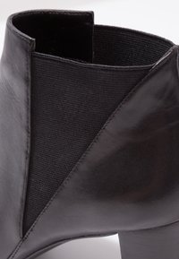 Gros plan sur une botte cheville en cuir noir avec panneau latéral élastique et petit talon bloc sur un fond uni.