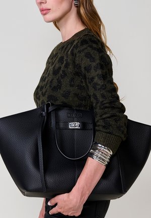 Gran bolso tote de cuero negro con textura granulada, que presenta herrajes plateados y un asa superior. Llevado por una persona con un suéter verde oliva.