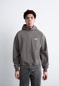 Grå hætte sweatshirt med lommer foran og logo på brystet, båret med grå jeans. Stoffet virker blødt med en afslappet pasform.