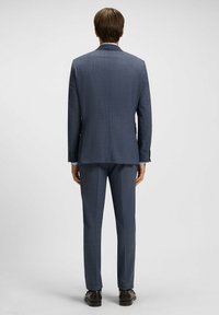 Marineblauwe suit met een gestructureerde stof, colbert met één rij knopen, slim-fit broek en zwarte leren schoenen, gezien vanuit de achterkant.