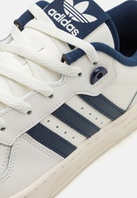 Sneaker in pelle bianca con dettagli blu navy, design a tre strisce, suola in gomma texturizzata e linguetta in tessuto con logo.