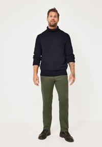 Dunkelblau gestrickter Rollkragenpullover kombiniert mit olivgrünen Hosen und schwarzen Schnürschuhen. Der Pullover hat einen gerippten Saum und gerippte Bündchen.