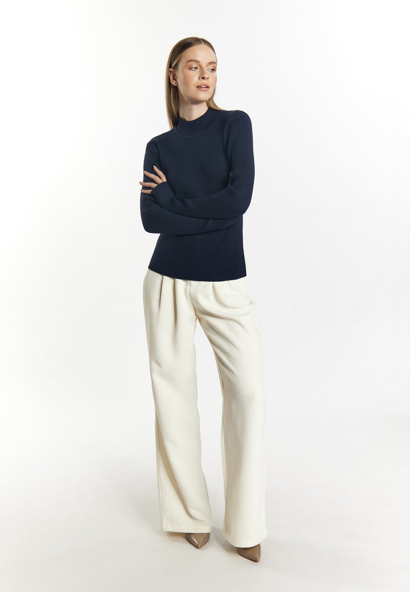 Marineblauwe geribbelde top met lange mouwen en hoge hals, gecombineerd met witte wijde broek en beige puntige hakken, tegen een lichte achtergrond.